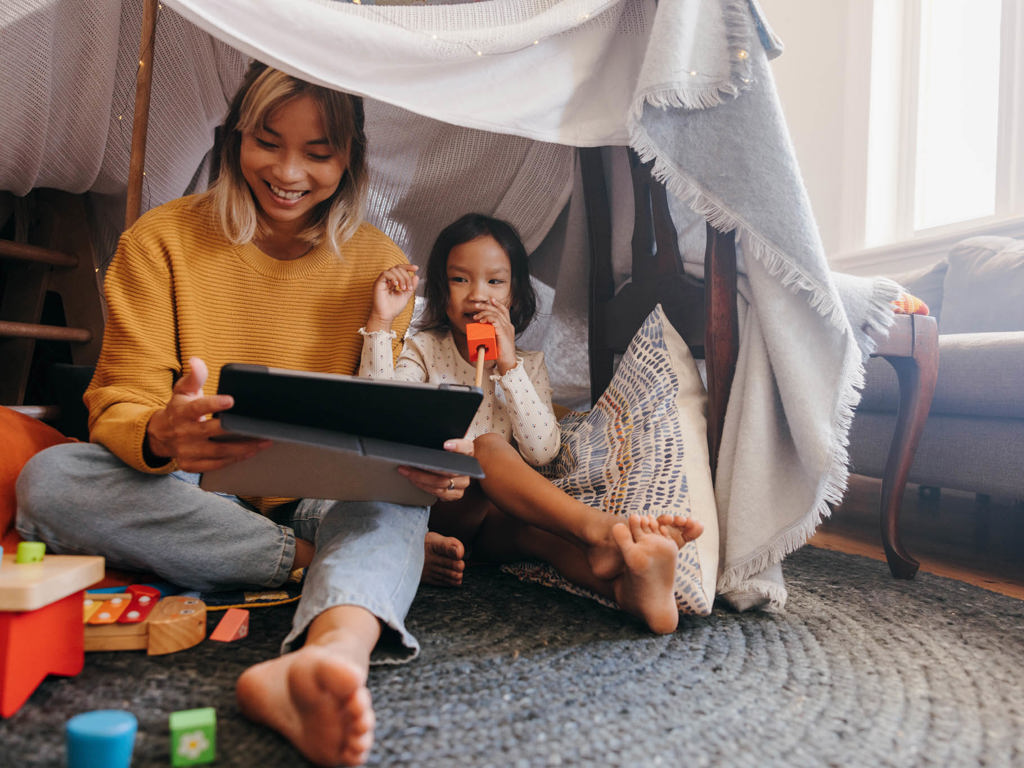 En mor og datter sidder i et hjemmelavet telt og kigger på en tablet