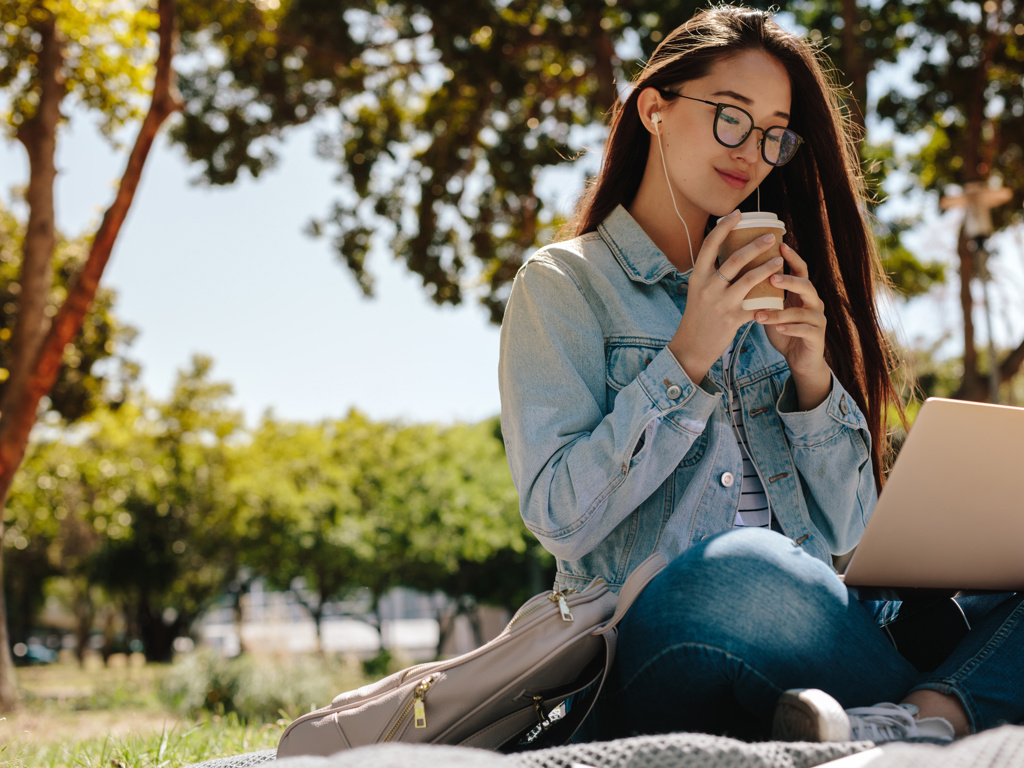 En kvinder sidder i en park med sin bærbar og høre telefoner, imens hun drikker kaffe